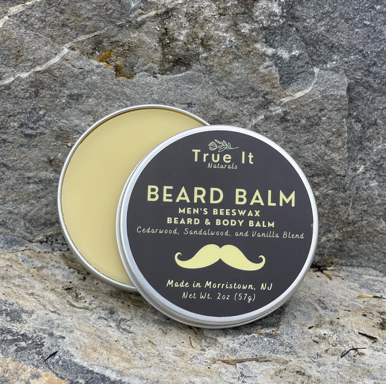 True It Naturals Beard Balm container with the lid open on a stone surface showing the beeswax salve. 