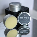 Bee Basic unscented beeswax lip and body balm tins on a reflective surface with a blurred background.