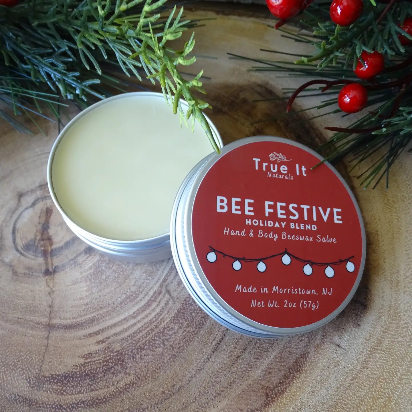 True It Naturals Bee Festive holiday blend beeswax salve with a red label on a wooden surface with greenery and berries.