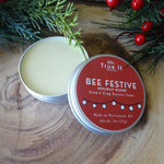 True It Naturals Bee Festive holiday blend beeswax salve with a red label on a wooden surface with greenery and berries.
