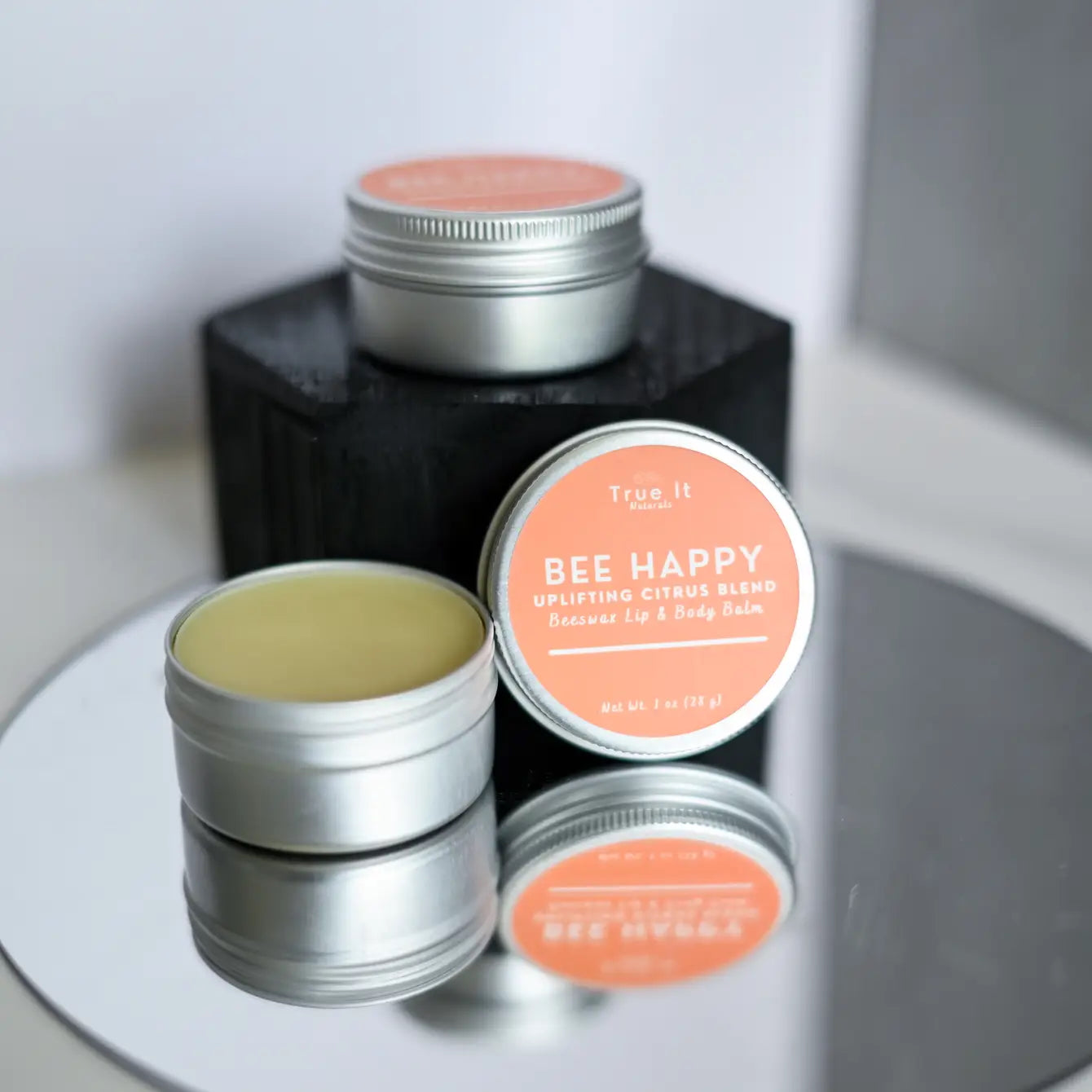 Two tins of True It Naturals Bee Happy Lip and Body beeswax balm on a reflective surface.