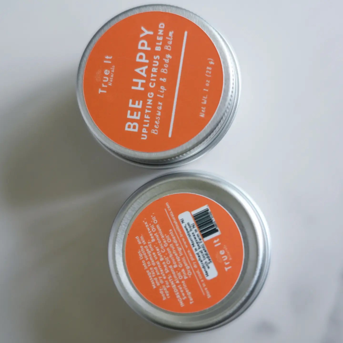 Two tins of True It Naturals Bee Happy beeswax lip and body balm showing front and back labels.