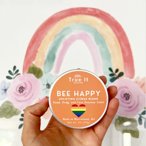 Hand holding a 'True It Naturals Bee Happy' beeswax salve tin with the special edition rainbow logo and a rainbow and floral background.