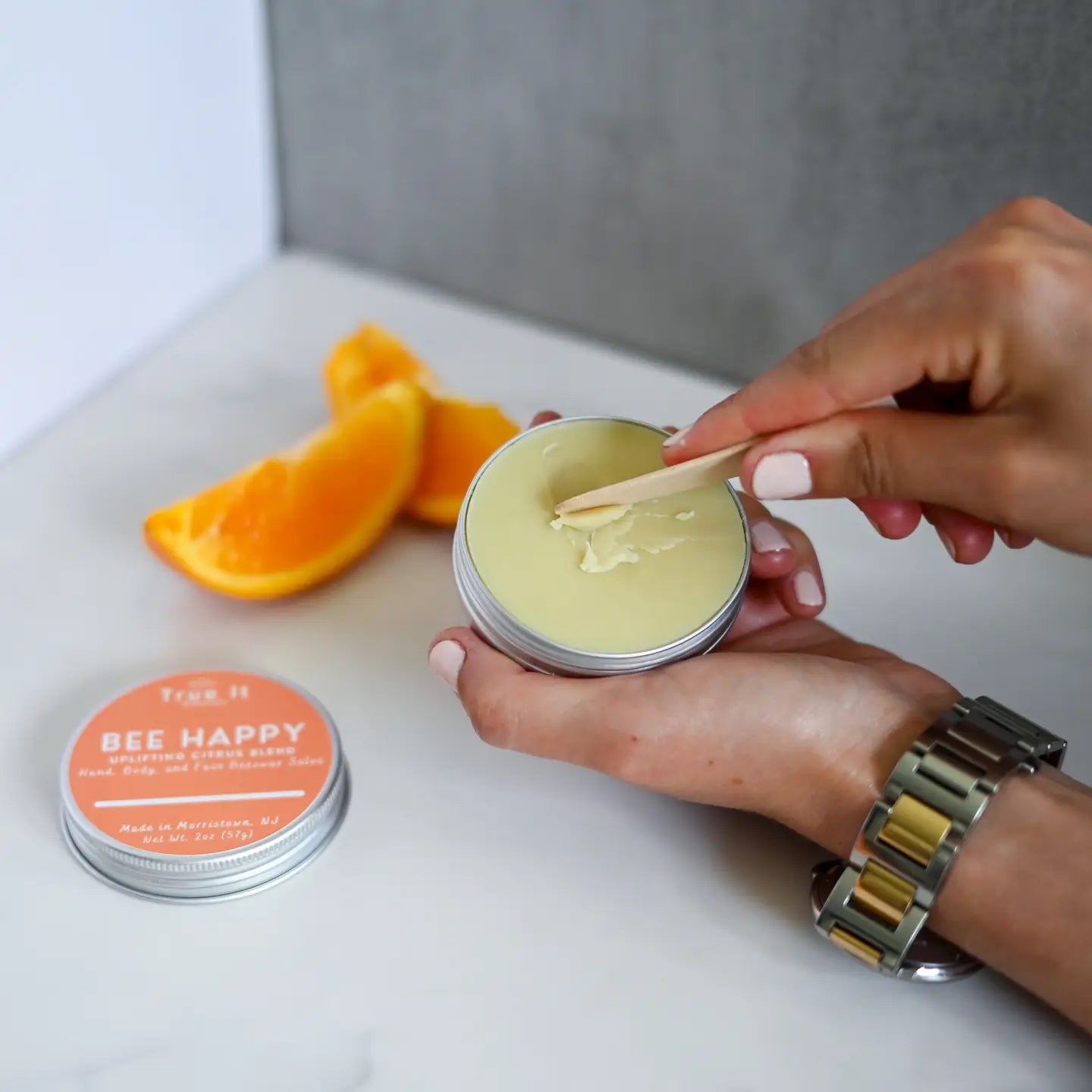 Person applying some 'Bee Happy' beeswax salve while holding the container with orange slices in the background on a white surface.