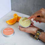 Load image into Gallery viewer, Person applying some &#39;Bee Happy&#39; beeswax salve while holding the container with orange slices in the background on a white surface.
