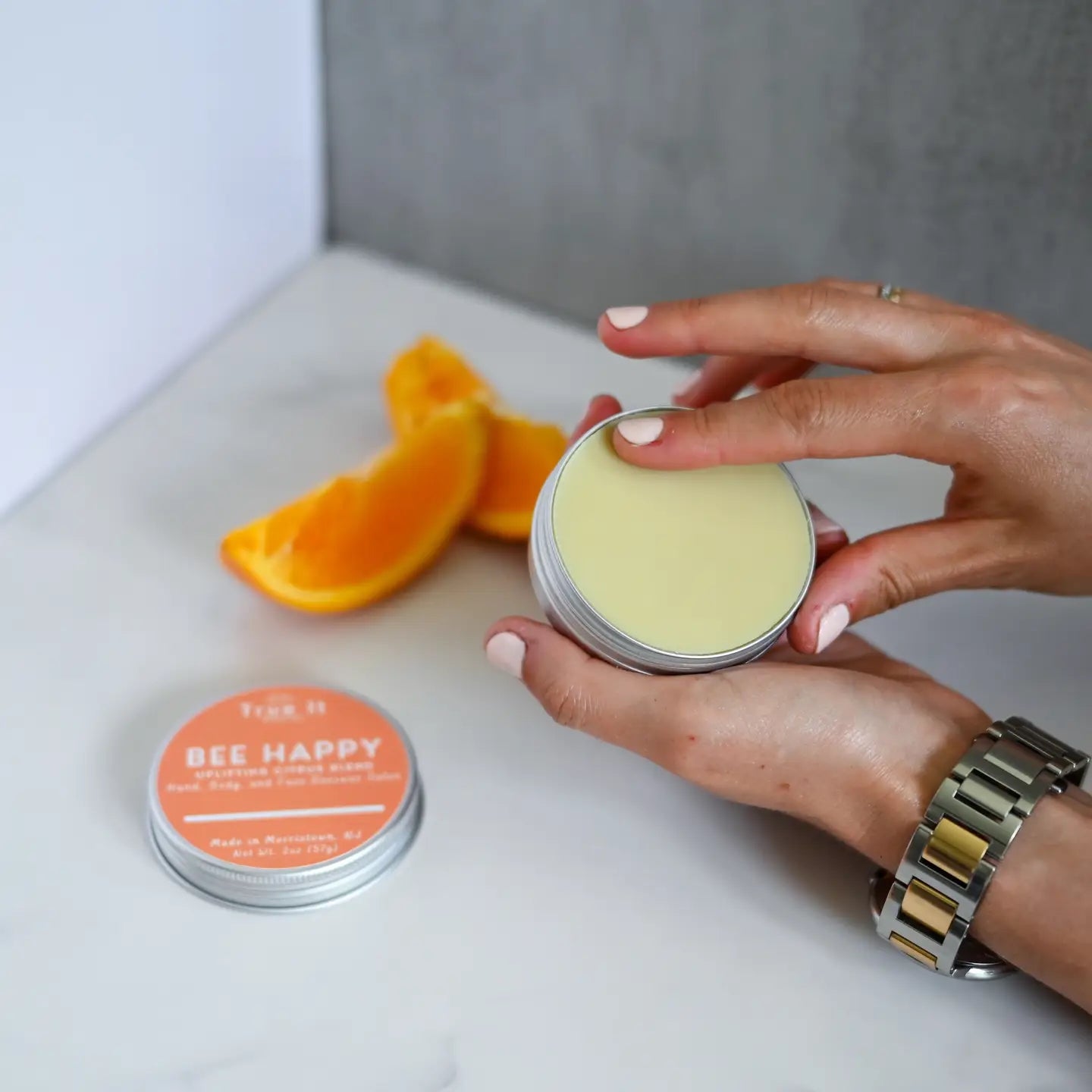 Hand holding a container of Bee Happy beeswax salve with another container and orange slices on a white surface