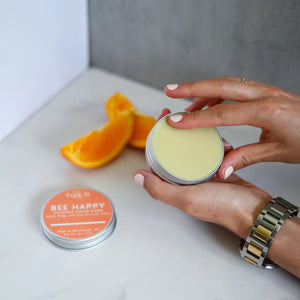 Hand holding a container of Bee Happy beeswax salve with another container and orange slices on a white surface