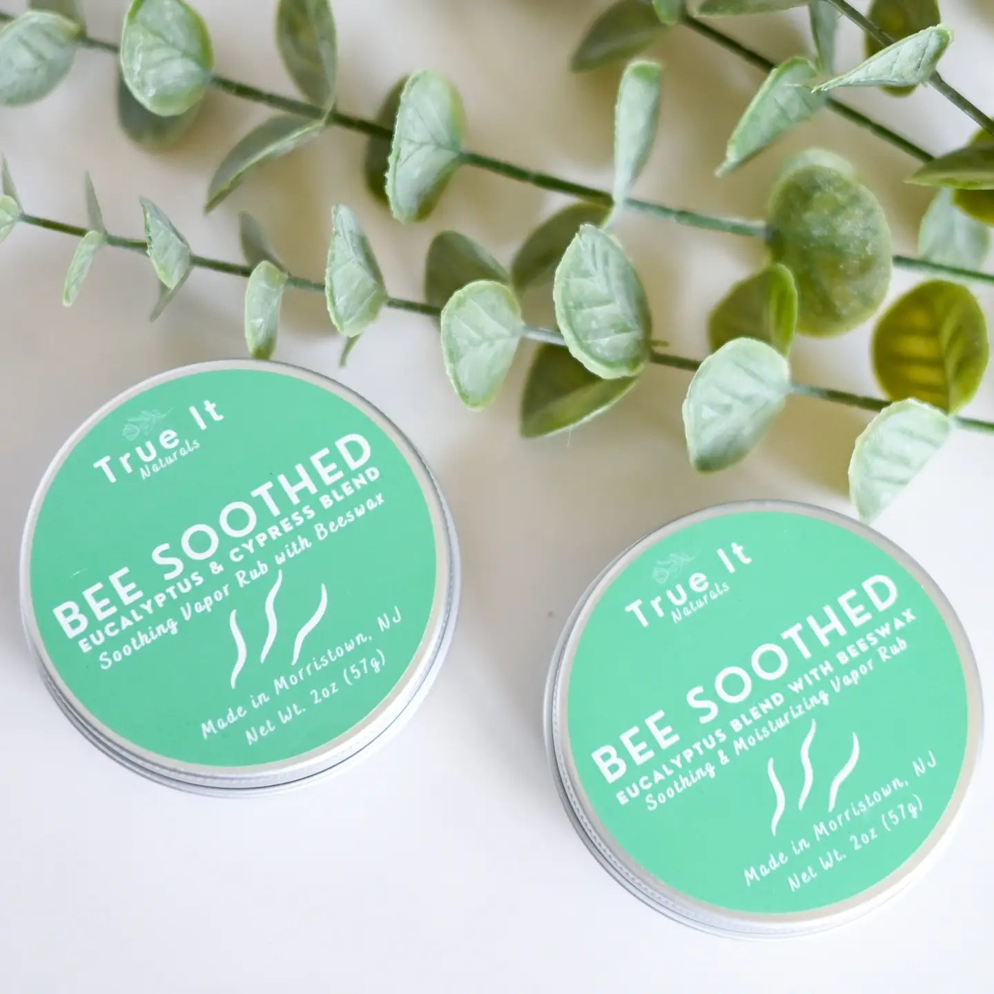 Two containers of 'True It' Bee Soothed beeswax salve on a white background with Eucalyptus leaves.
