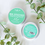 Two containers of 'True It Bee Soothed' beeswax salve with green labels on a light background with Eucalyptus leaves.