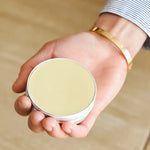 Load image into Gallery viewer, Hand holding a 2 oz beeswax salve on a beige background
