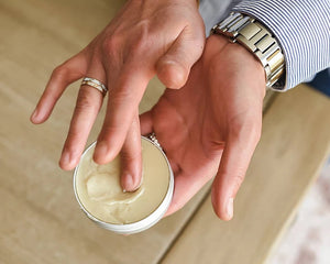 Hand applying cream from a small container on a neutral background