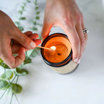 Load image into Gallery viewer, Person lighting a True It Naturals Beeswax Candle - Vanilla Sage | 100% Beeswax, inside an amber glass jar with a wooden wick, on a light background.
