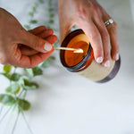Person lighting a True It Naturals Beeswax Candle - Vanilla Sage | 100% Beeswax, inside an amber glass jar with a wooden wick, on a light background.