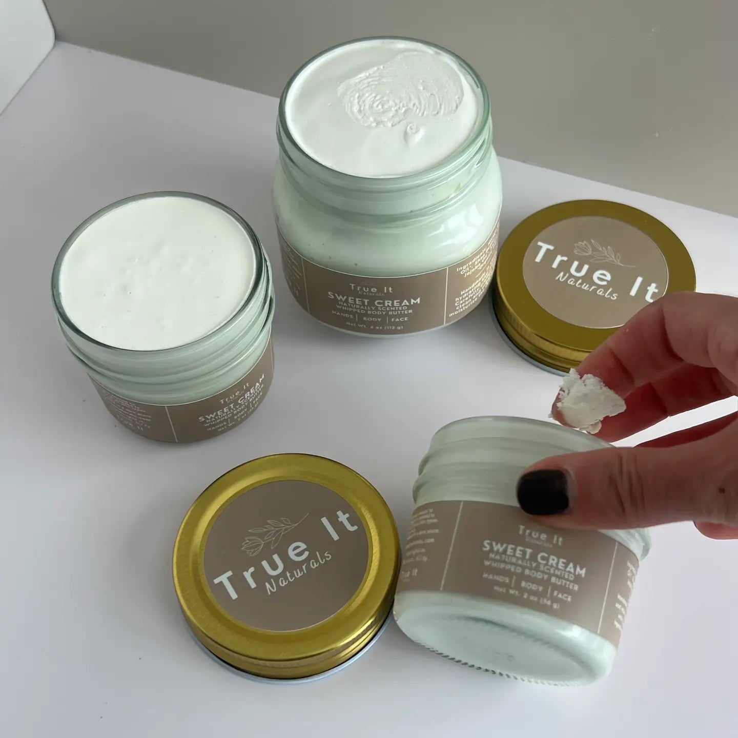 Jars of Small (2oz) and Large (4oz) True It Naturals Sweet Cream with the lids off showing the cream texture. Also someone scooping some cream out of a jar with a finger.