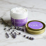 True It Naturals small jar of Lavender Cream with lid off and lavender flowers on a marble surface